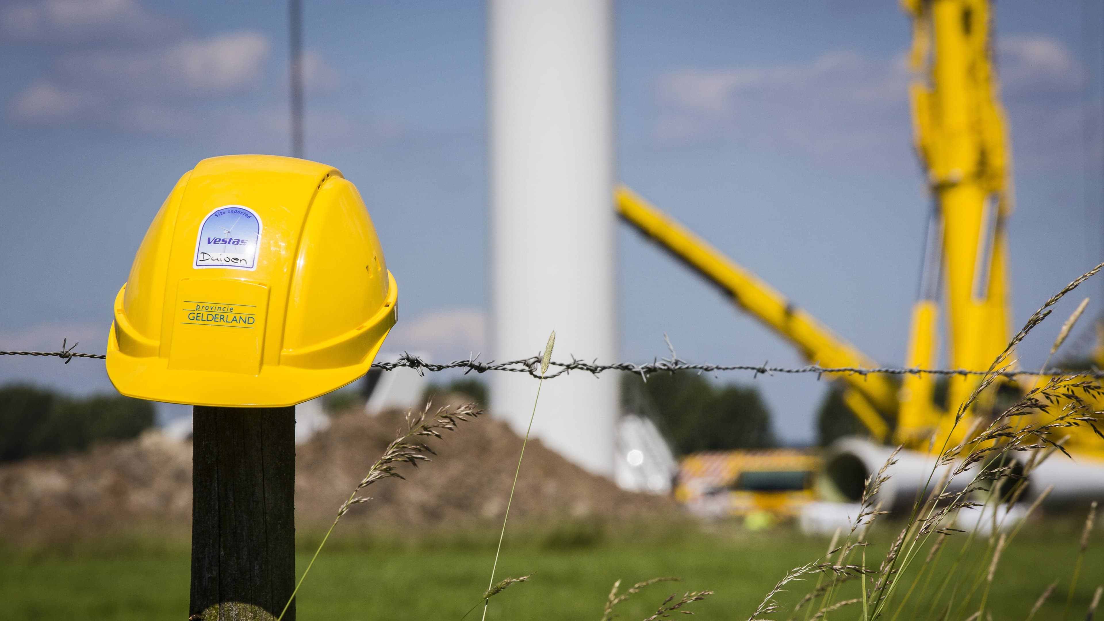 Gele bouwhelm op een paaltje van een hek met op de achtergrond een windturbine in aanbouw